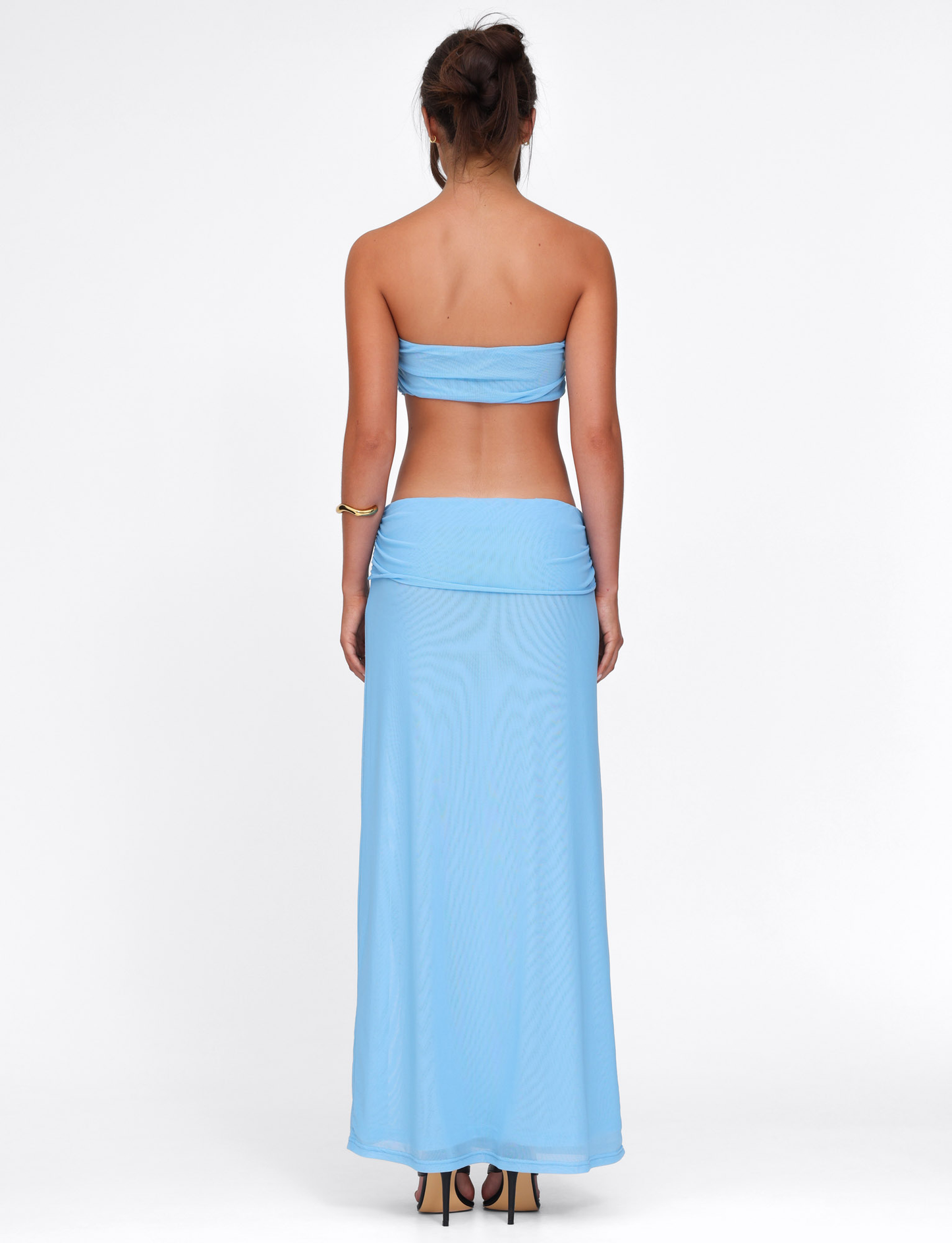 A woman with an updo wears the TIGERMIST KYNLEE DRESS in sky blue, featuring a strapless neckline and matching skirt. She accessorizes with a gold bracelet and stands against a plain white background.